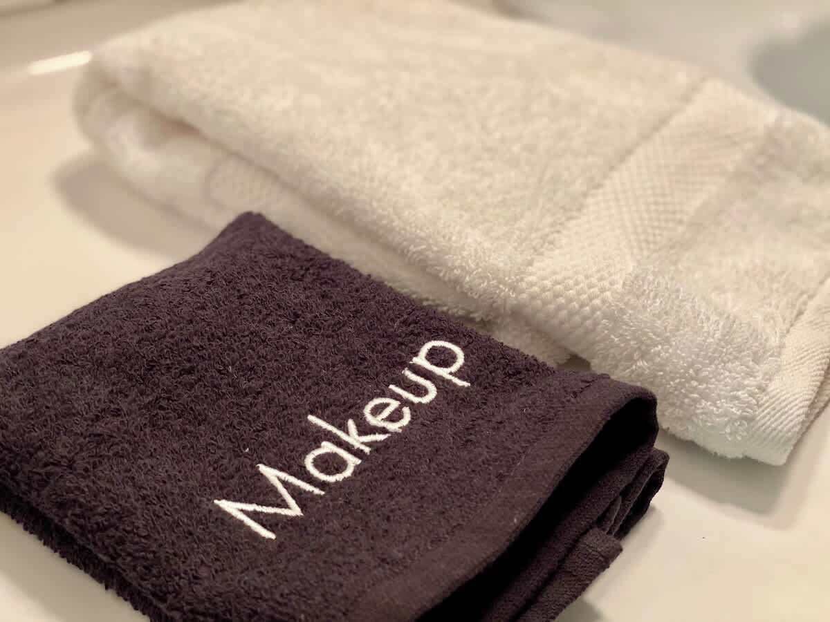 Une serviette pliée de couleur marron foncé, ornée du mot « Maquillage » brodé dessus, et une serviette blanche à côté, posées sur une surface blanche.