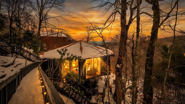 Ein mit Schnee bedecktes Haus mit einer Holzdielen-Terrasse und warmem Licht im Inneren.