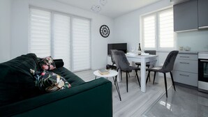 Un salon moderne avec un canapé vert, un coin repas avec une table et des chaises blanches, et une cuisine avec des meubles gris.