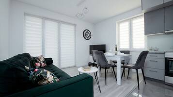 Una sala de estar moderna con un sofá verde, una zona de comedor con una mesa y sillas blancas, y una cocina con armarios grises.