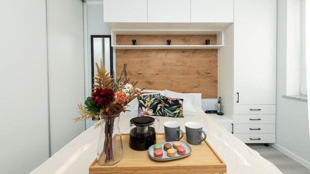 Une chambre à coucher moderne avec une tête de lit en bois, une table de chevet sur laquelle se trouvent une cafetière, des tasses et une assiette de macarons.