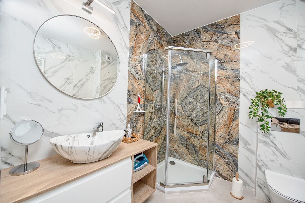 Une salle de bain moderne avec un lavabo en marbre, un miroir rond, une douche en verre et une plante en pot.