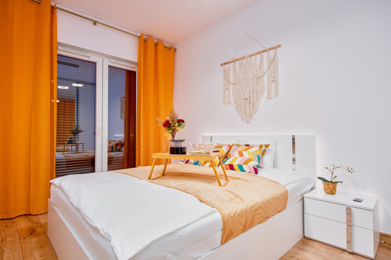 Um quarto com cama, mesa de cabeceira, luminária e um pendente de macramé na parede, além de uma janela com cortinas.
