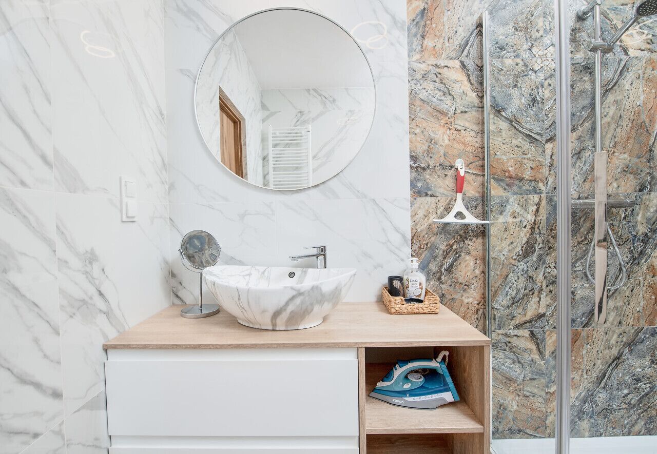 Een badkamer met een marmeren wand, een ronde spiegel, een witte wastafel en een douchegedeelte.