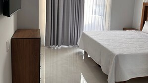 Una habitación de hotel moderna con una cama grande, una mesita de noche de madera, un televisor de pantalla plana montado en la pared y un ventanal con cortinas grises.