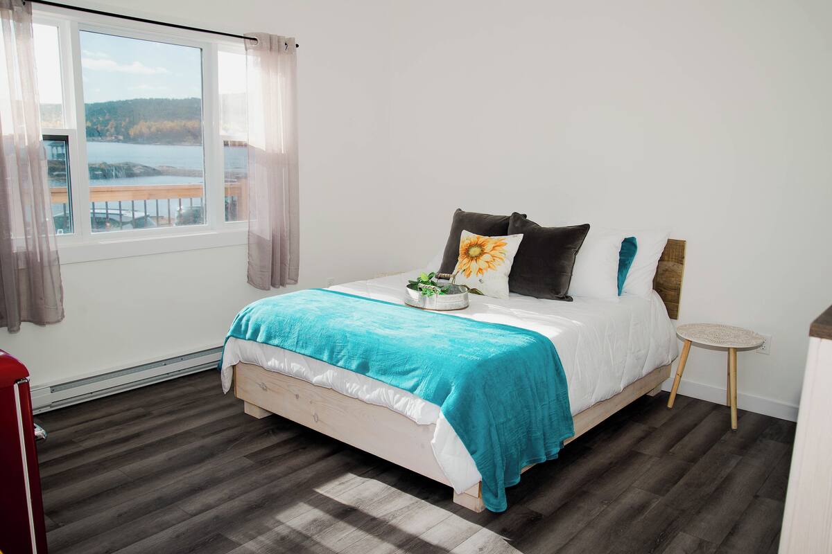 A bedroom with a large window, a bed with a teal blanket, and a small round table.