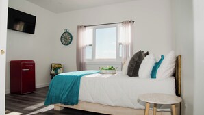 Une chambre à coucher avec un lit, une petite table, un appareil électroménager rouge, une horloge et une fenêtre avec des rideaux.