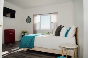 Un dormitorio con cama, una mesita, un electrodoméstico rojo, un reloj y una ventana con cortinas.