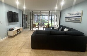 Una sala de estar con un sofá negro, un televisor montado en la pared y un ventanal corredero que da a un espacio al aire libre.