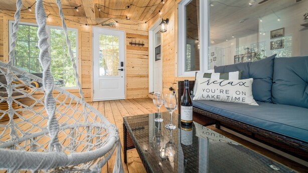 En hyggelig hyttekrog med en hængekøje, et glasbord med vinglas og en vinflaske, samt en sofa med pude, der står 'slap af, du er ved søen'.