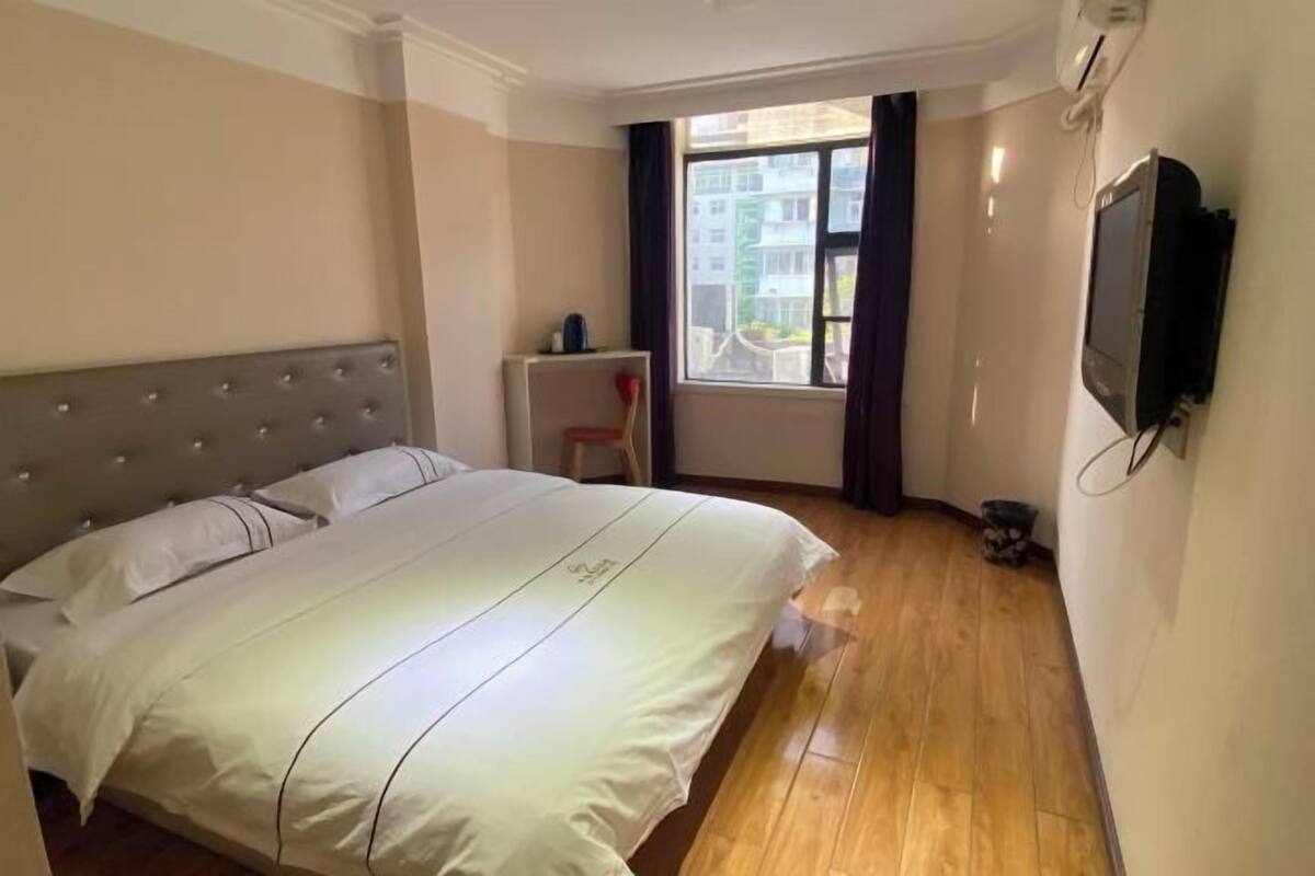 Hotellihuone, jossa on suuri sänky, pieni pöytä, tuoli, ikkuna verhoilla ja seinälle asennettu televisio.