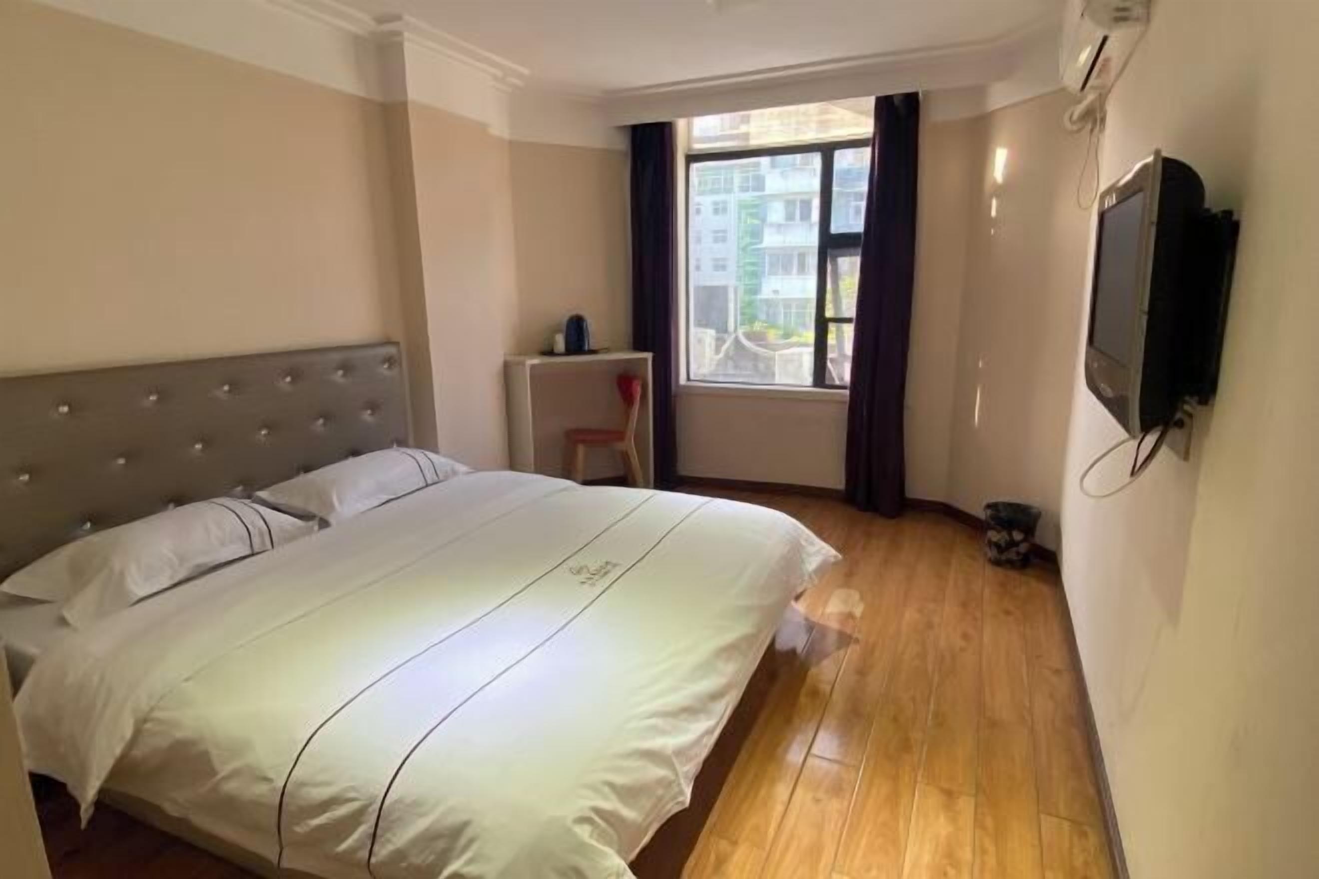 Habitación de hotel con una cama grande, una mesita pequeña, una silla, un televisor montado en la pared y un ventanal con cortinas.