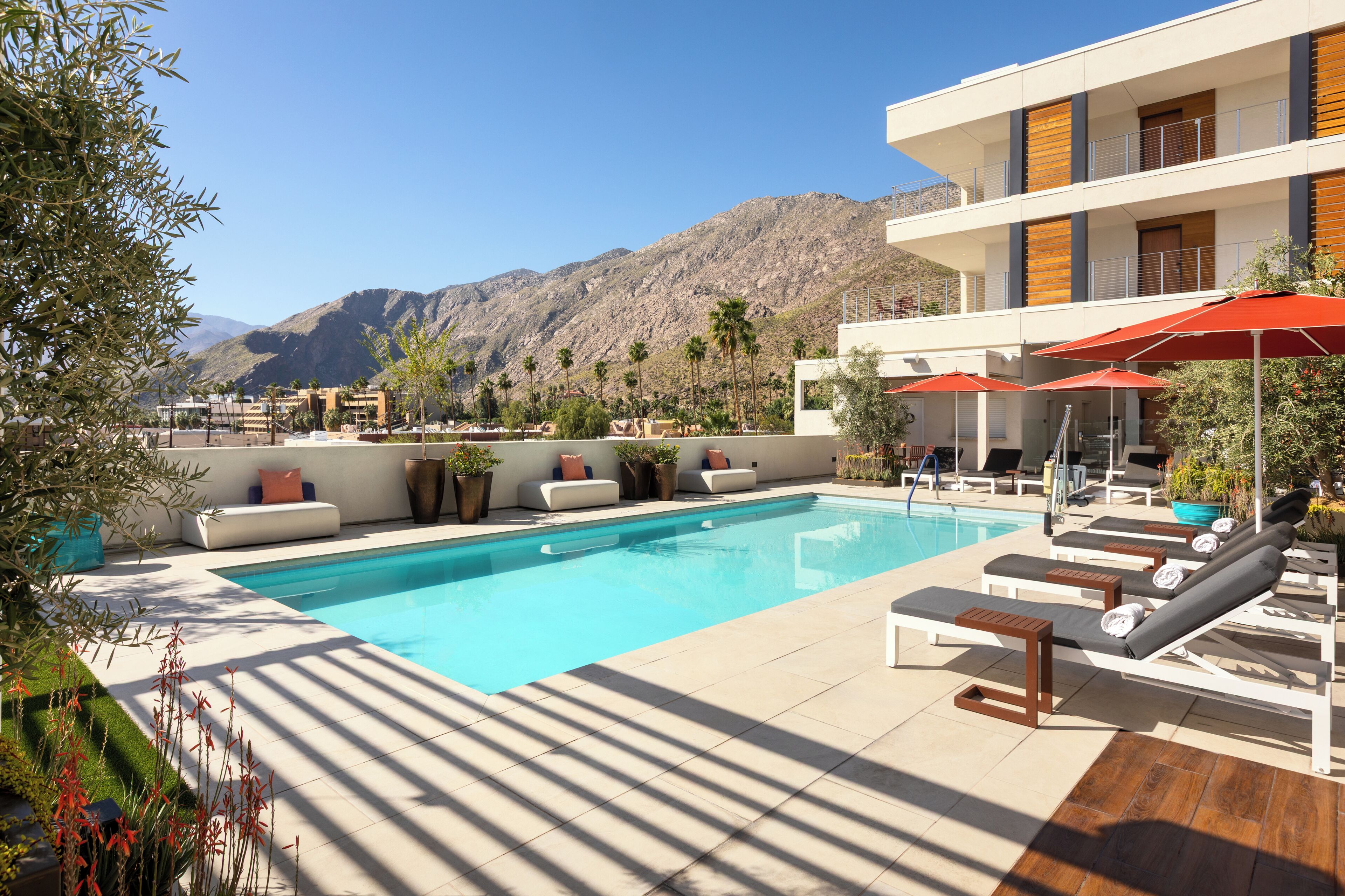 Un área de piscina con sillones de descanso y sombrillas, un edificio blanco con detalles de madera, y montañas al fondo.