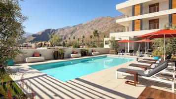 Un área de piscina con sillones de descanso y sombrillas, un edificio blanco con detalles de madera, y montañas al fondo.