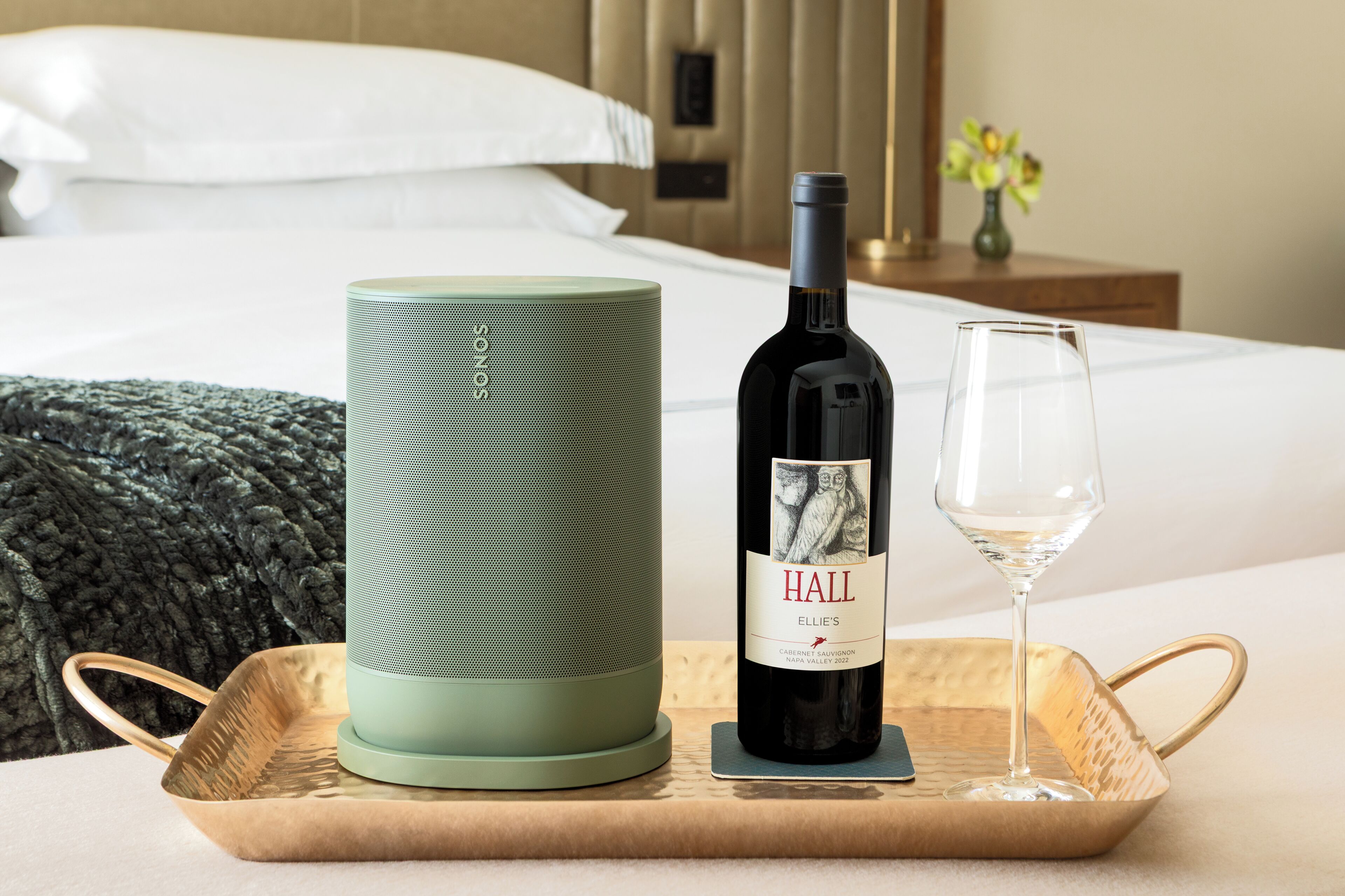 Una cama con sábanas blancas, un altavoz cilíndrico verde, una botella de vino con la etiqueta 'HALL' sobre una bandeja y una copa de vino.