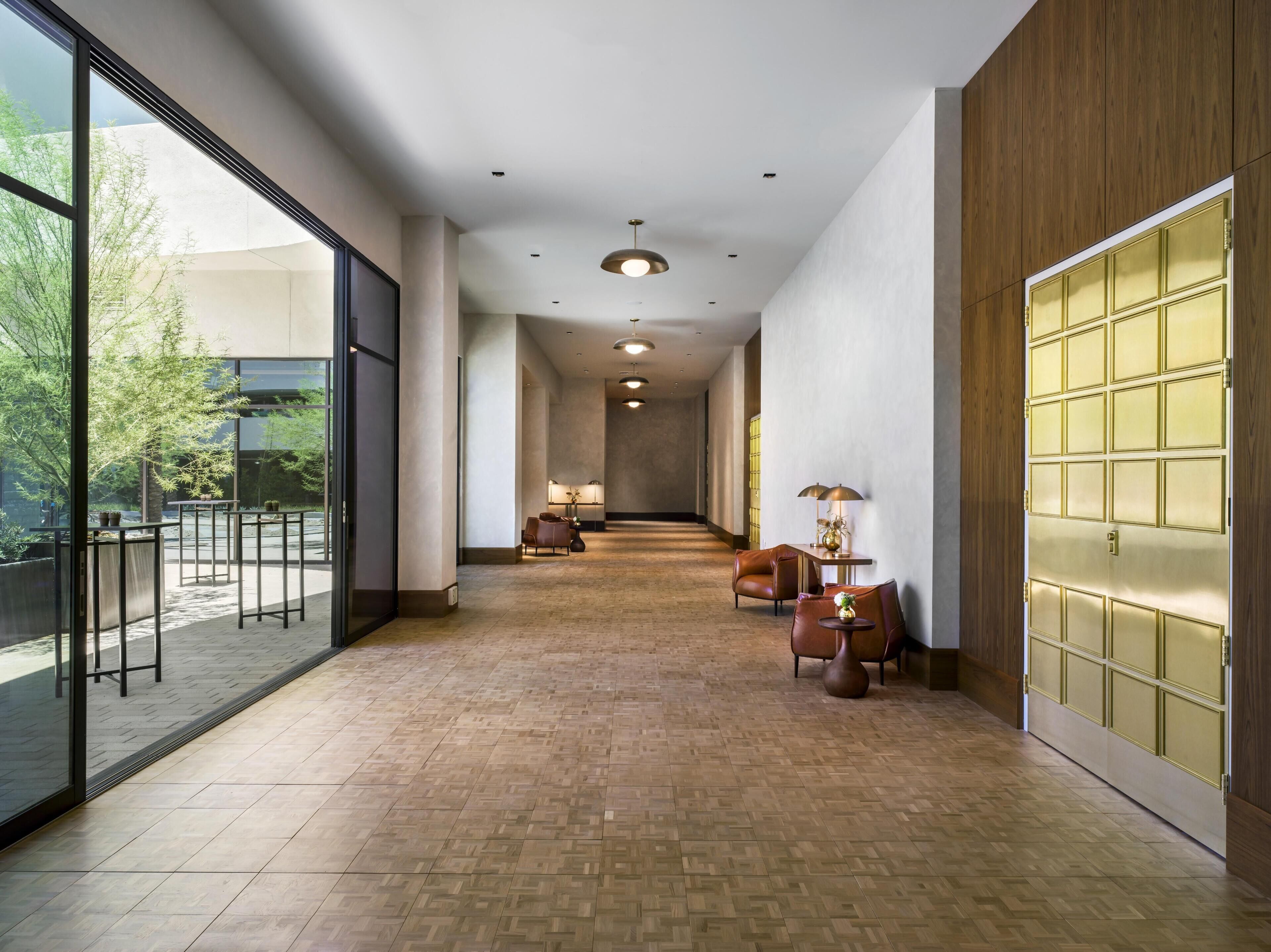 A modern hotel corridor with a wooden floor, glass doors, and a reception desk with chairs.