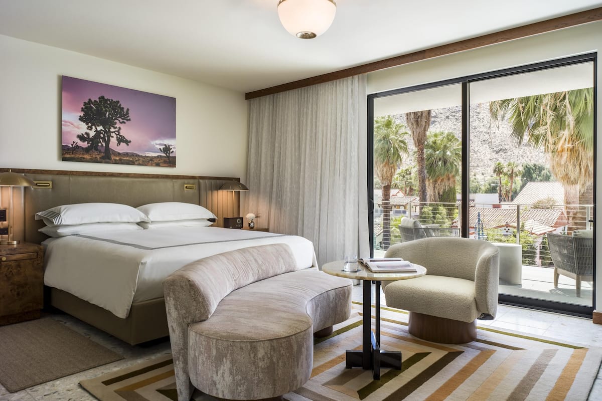 A hotel room with a bed, two armchairs, a small table, a painting, a lamp, and a view of palm trees through large windows.