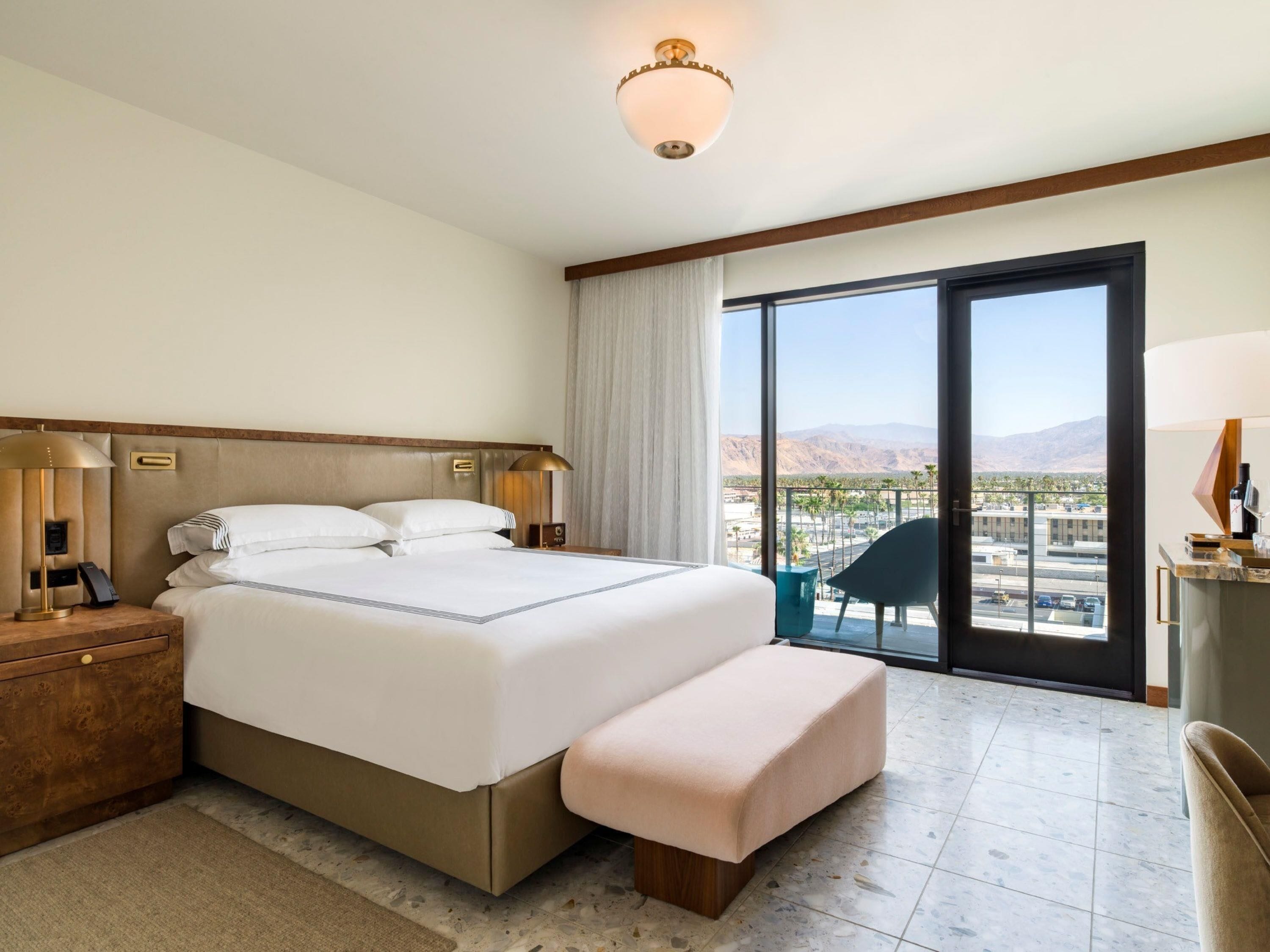 A hotel room with a large bed, a bedside table with a lamp, a chair by the window, and a view of the mountains.