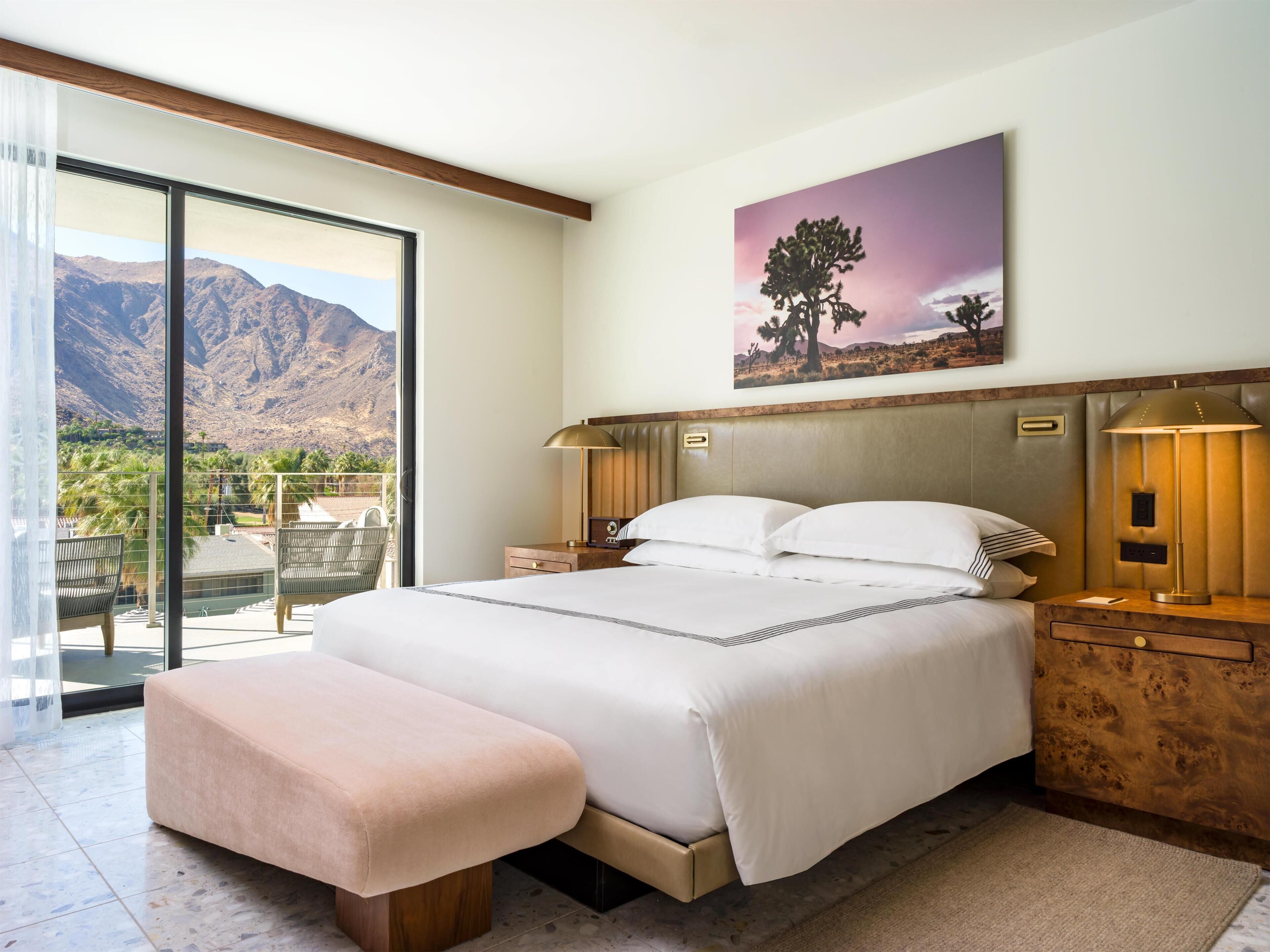 A bedroom with a large bed, bedside tables, a bench, a painting, and a view of mountains.