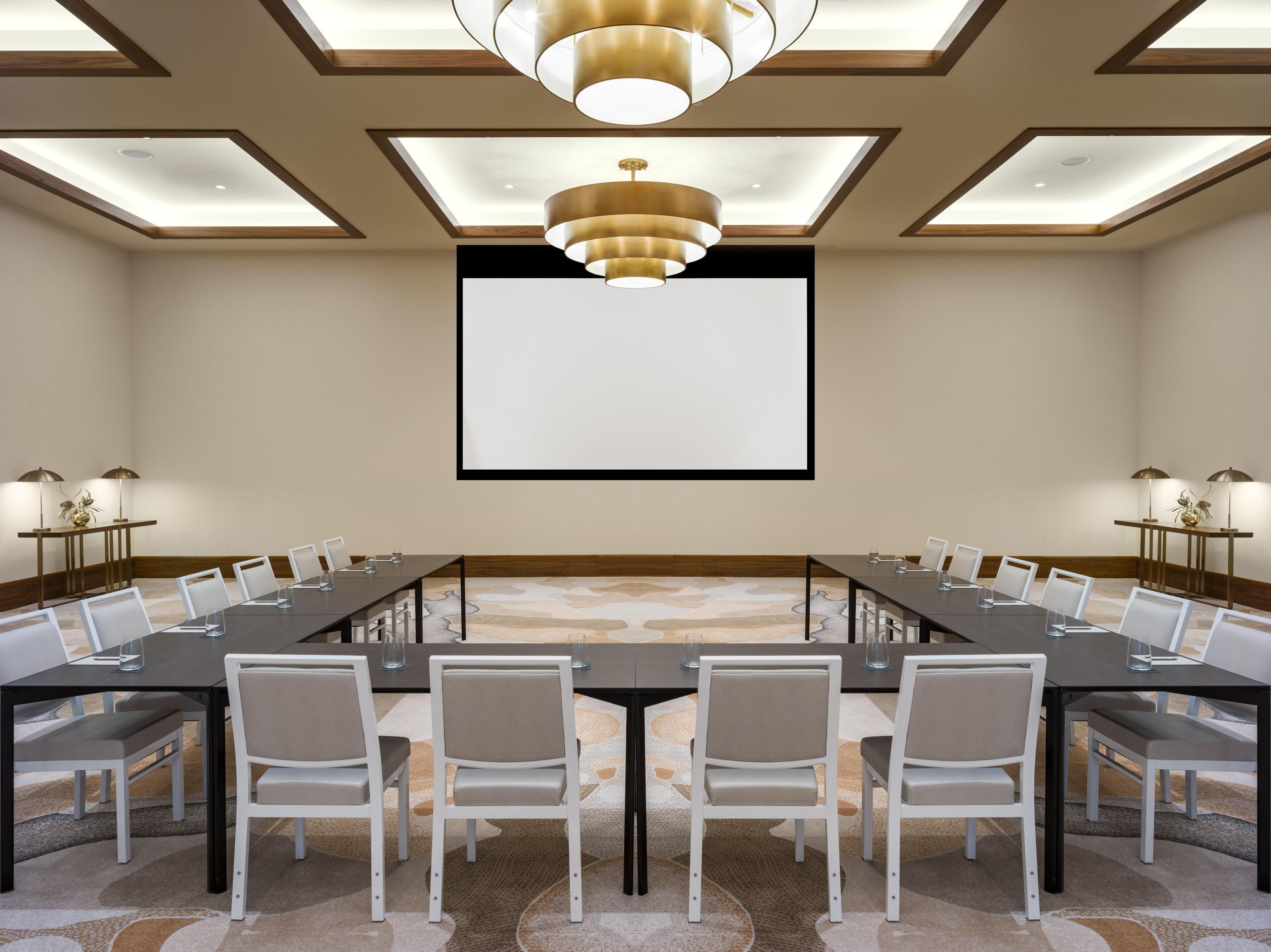 Un salón de conferencias con una larga mesa, sillas, una pantalla de proyector e iluminación colgante.