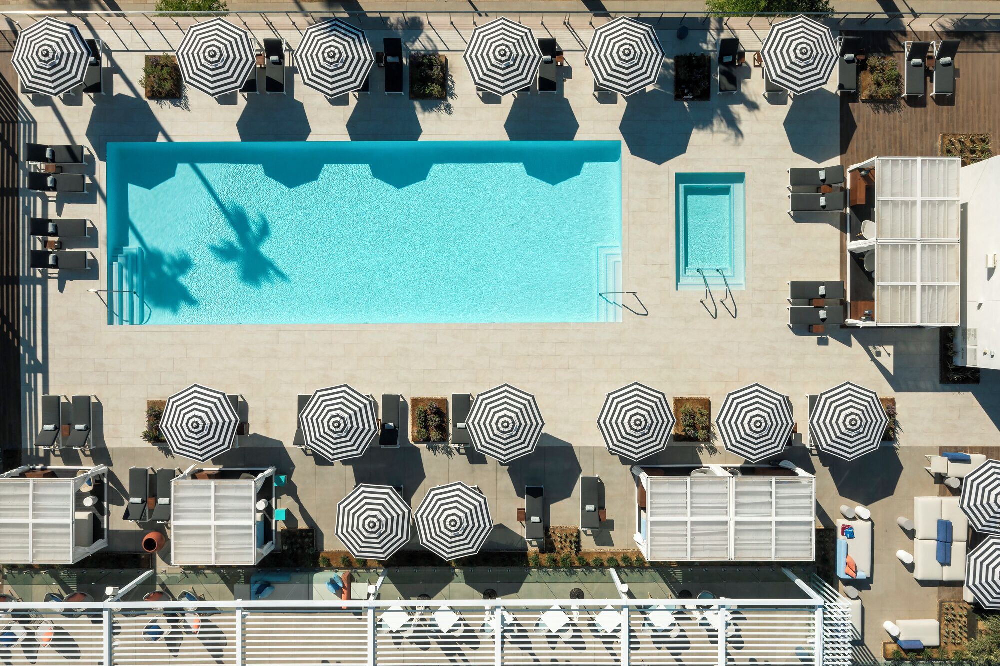 An aerial view of a hotel pool area with multiple lounge chairs and umbrellas, a large central pool, and a separate smaller pool.