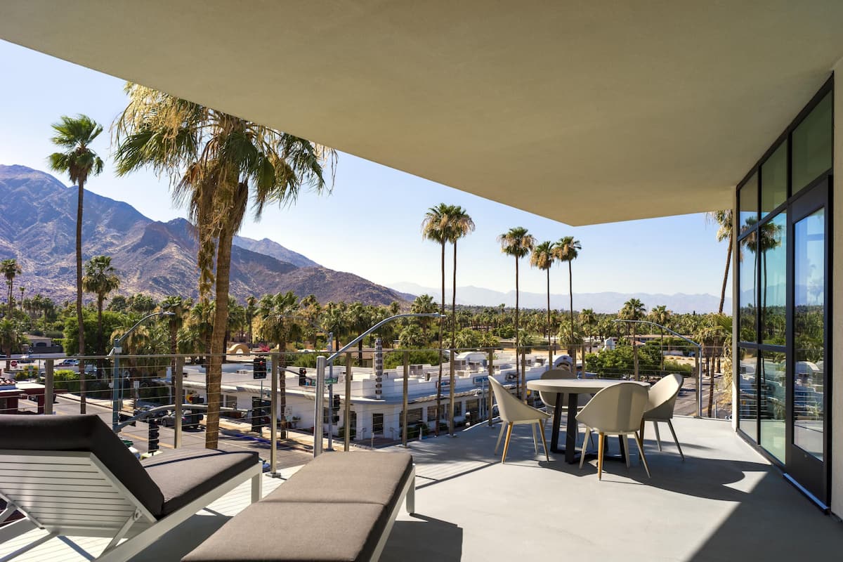 A balcony with lounge chairs, a table, and chairs, overlooking a parking area and palm trees with mountains in the background.