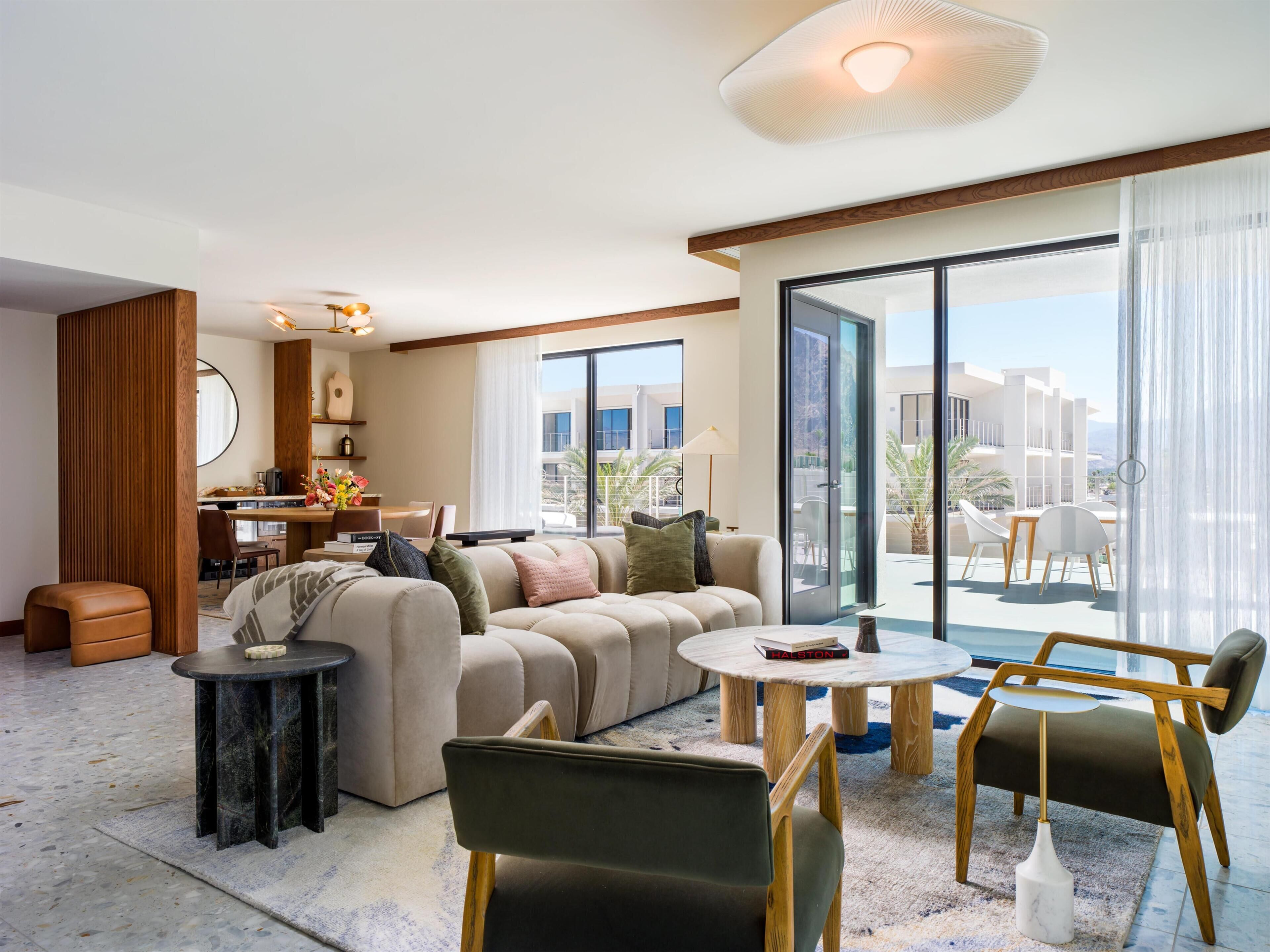 A modern hotel room with a sofa, armchairs, a dining area, and a balcony with a view.