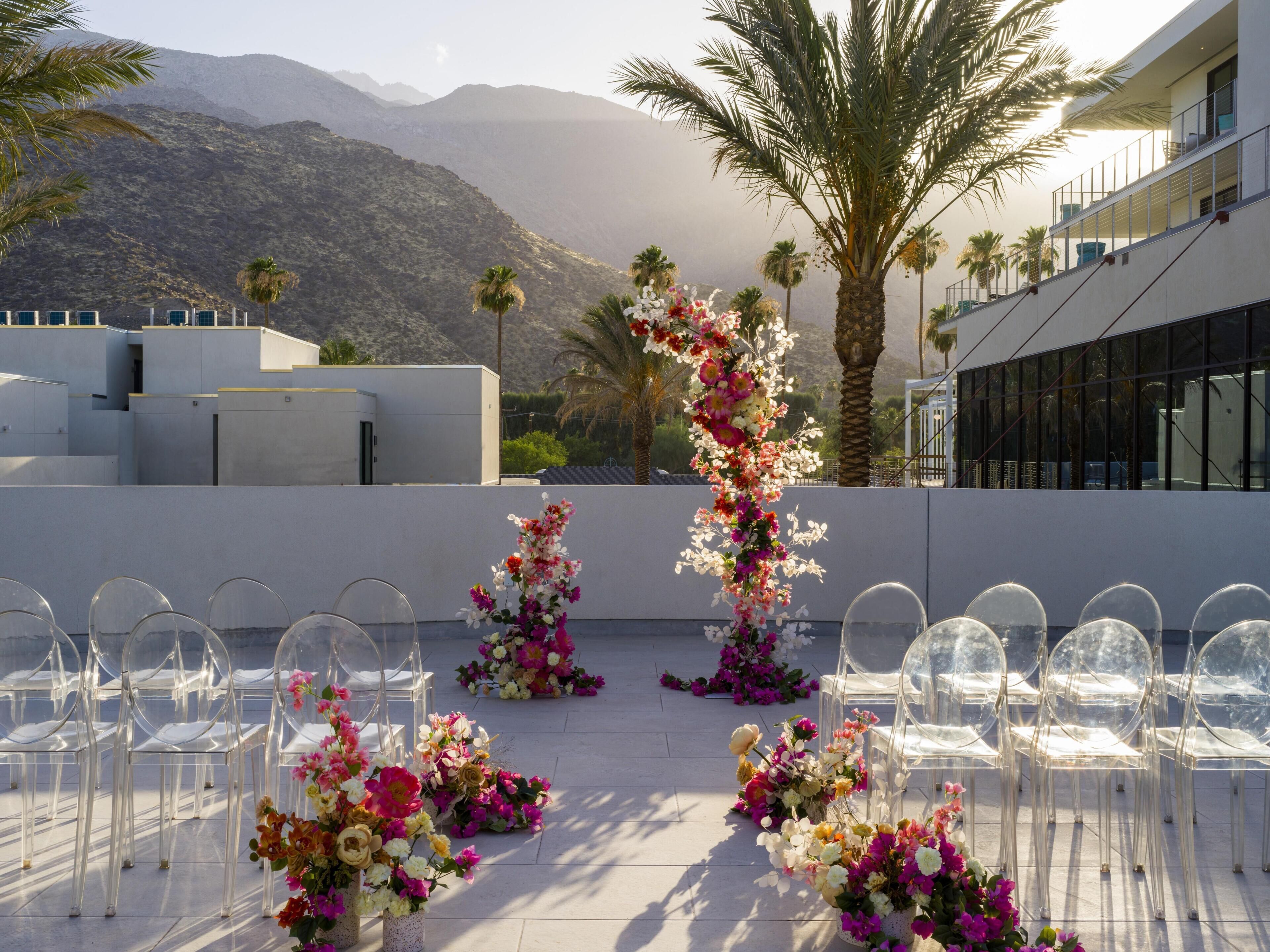 A rooftop venue with clear chairs arranged for an event, surrounded by floral decorations and palm trees, with a mountain backdrop.