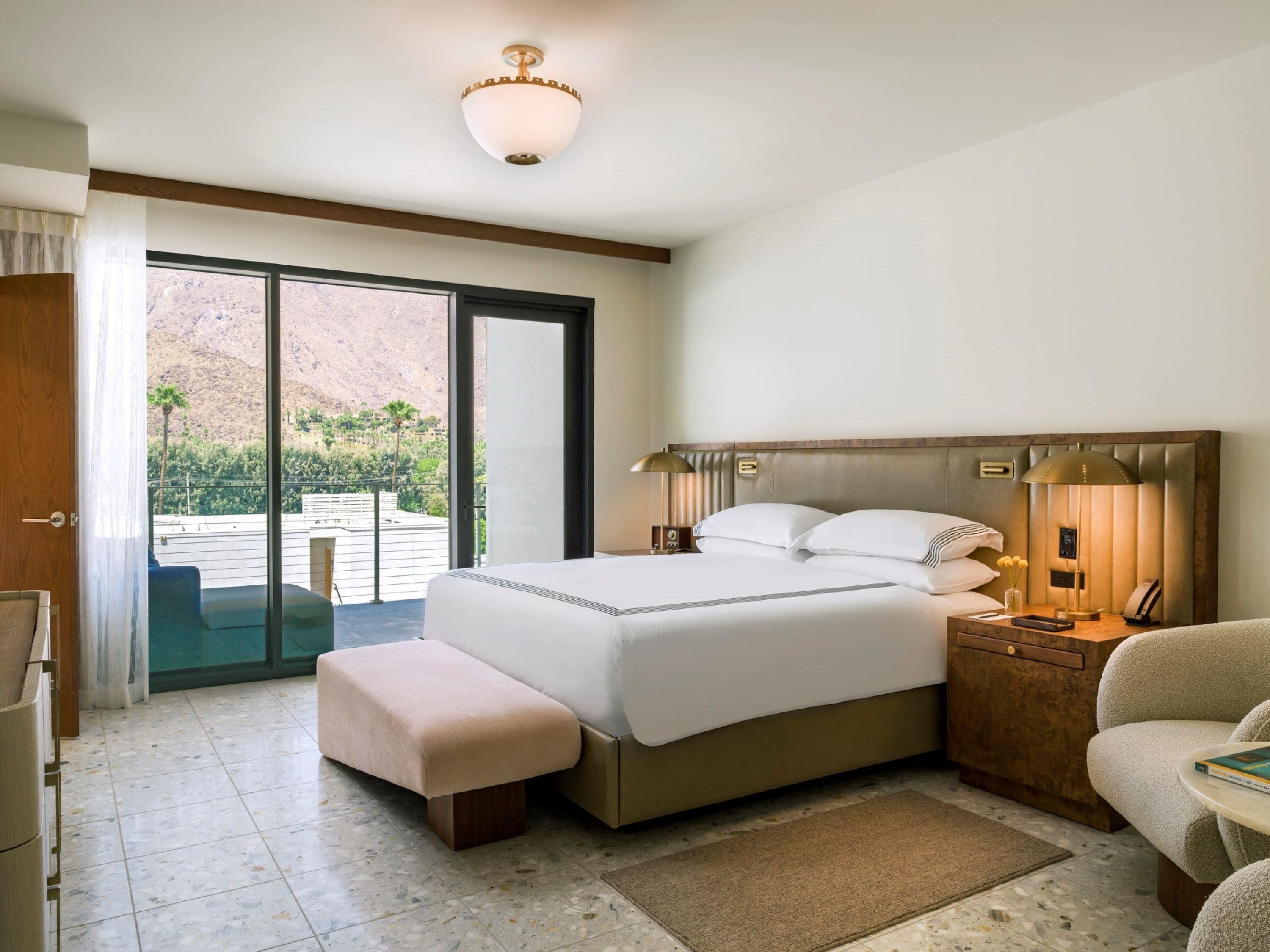 A hotel room with a bed, bedside table, lamp, and a view of a mountainous landscape through a sliding door.