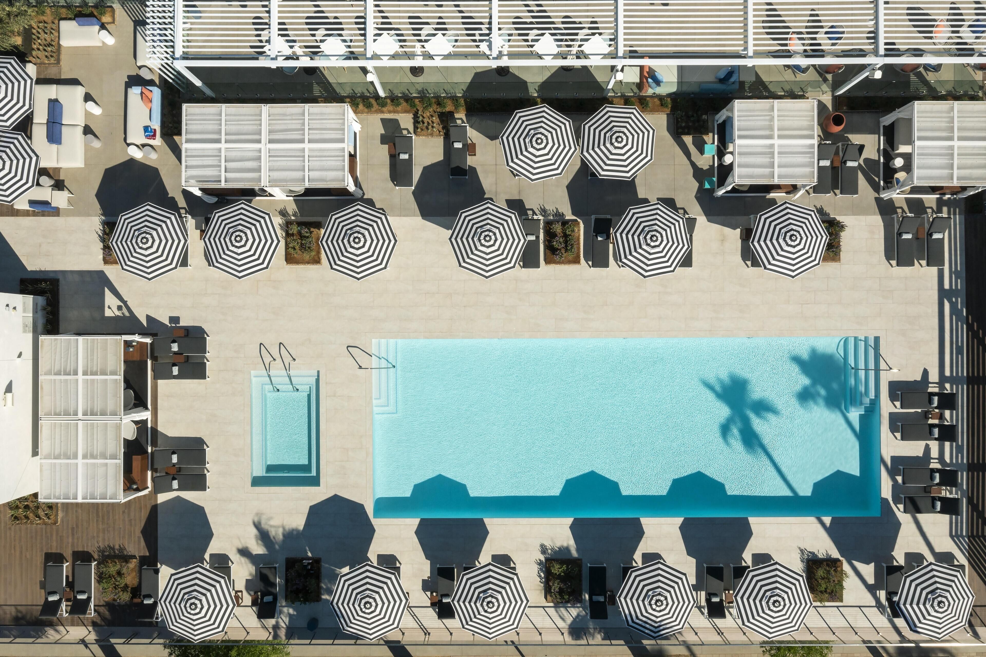 An aerial view of a rooftop pool area with multiple lounge chairs and umbrellas, a swimming pool, and a bar area.