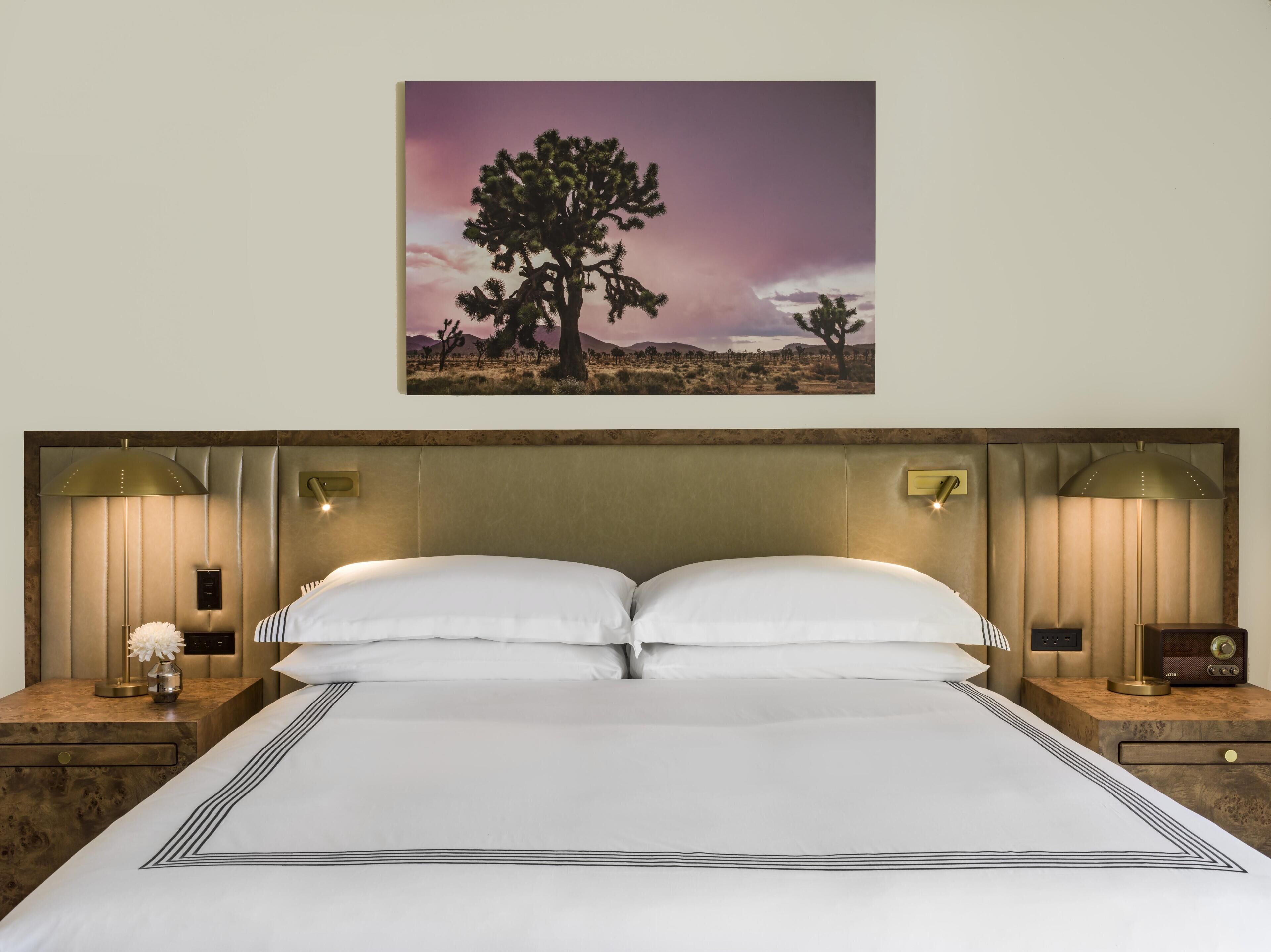 A neatly made bed with a headboard, two bedside lamps, a nightstand with a radio, and a framed picture of a desert landscape.