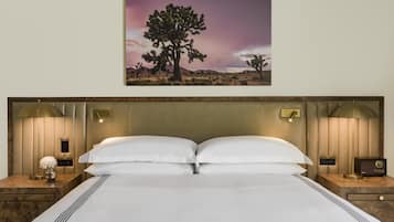 A neatly made bed with a headboard, two bedside lamps, a nightstand with a radio, and a framed picture of a desert landscape.