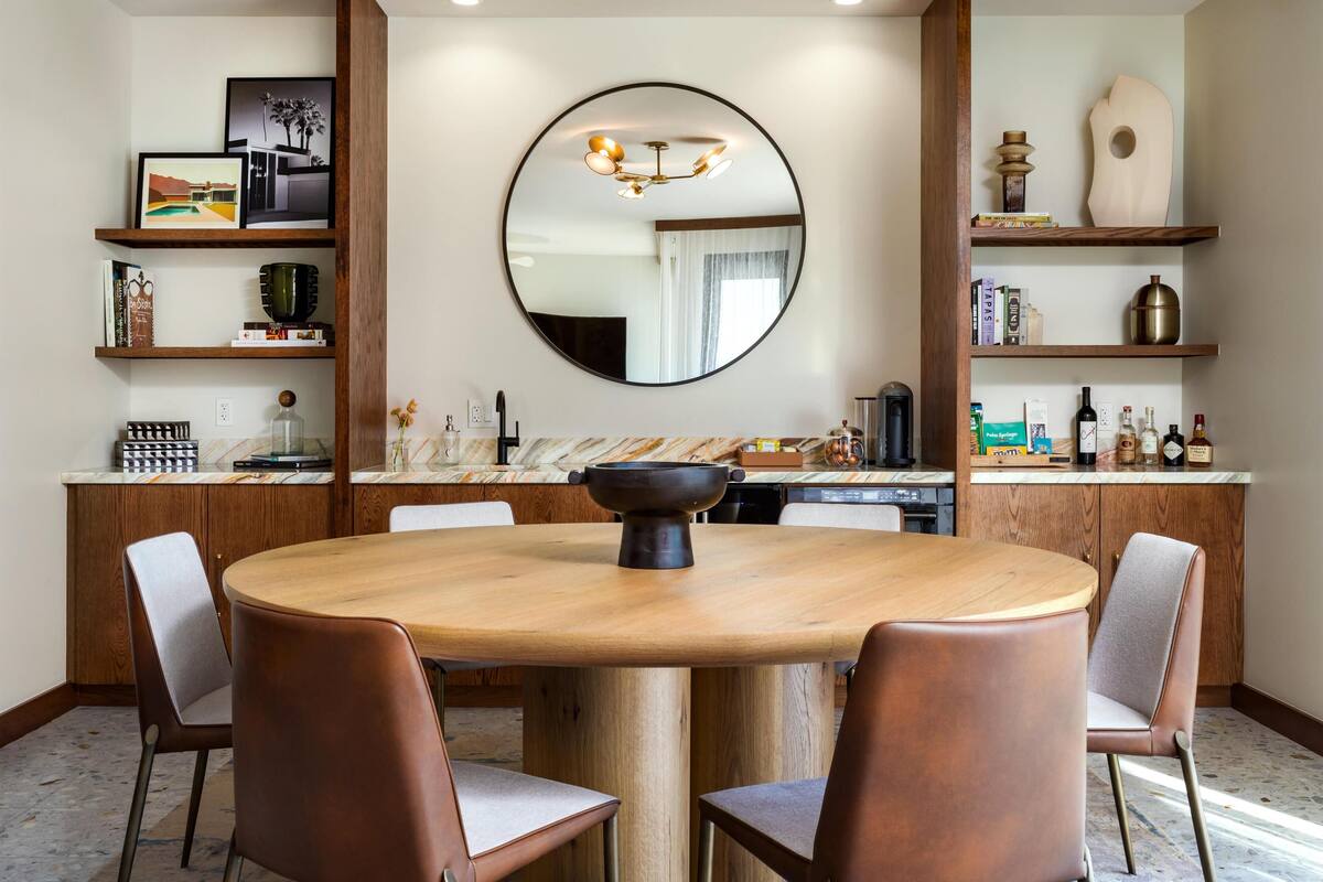 A dining area with a round table, six chairs, a central light fixture, and wooden shelves with books and decorative items.