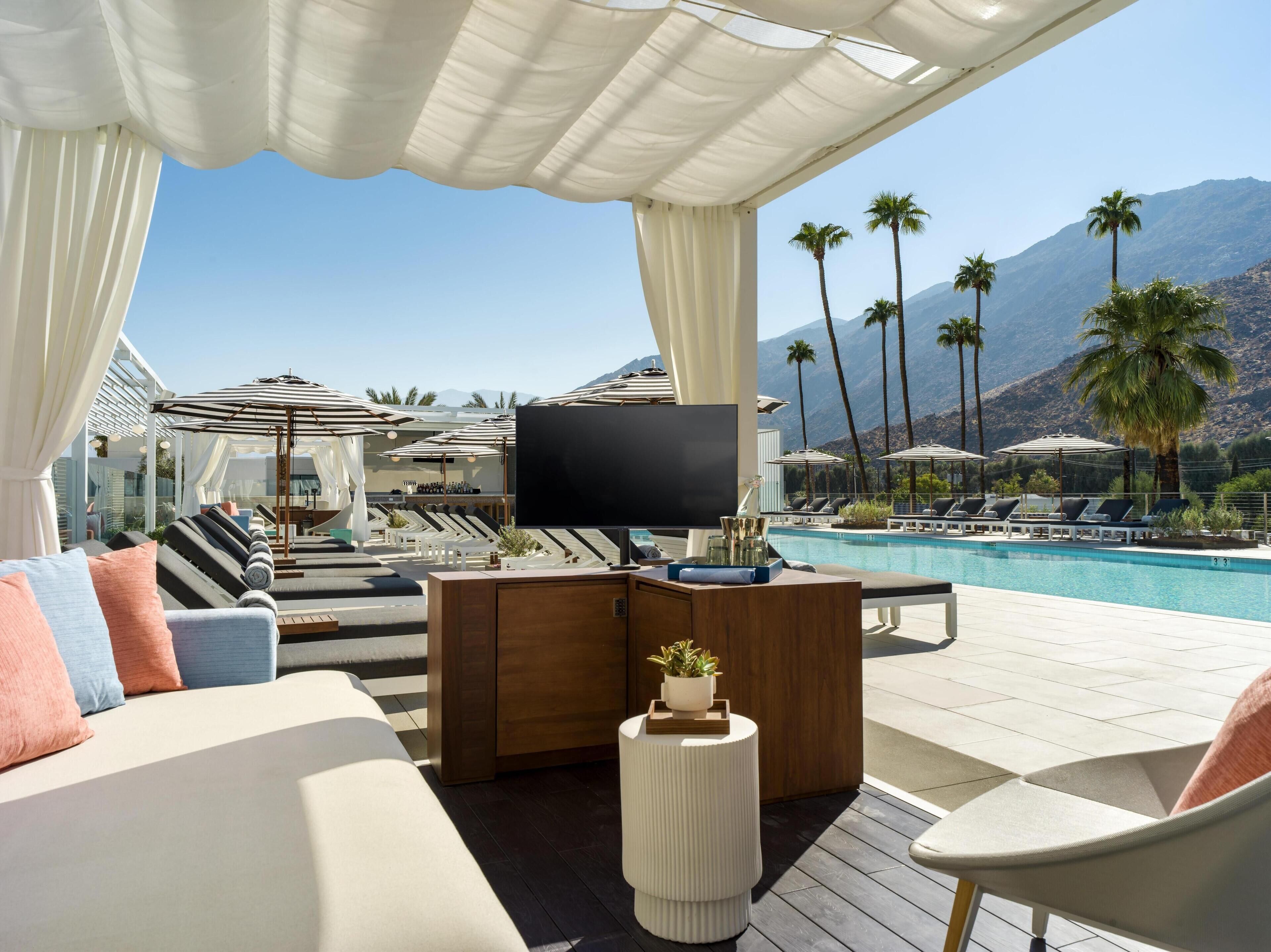 A poolside lounge with a flat-screen TV, cushioned seating, and a view of palm trees and mountains.