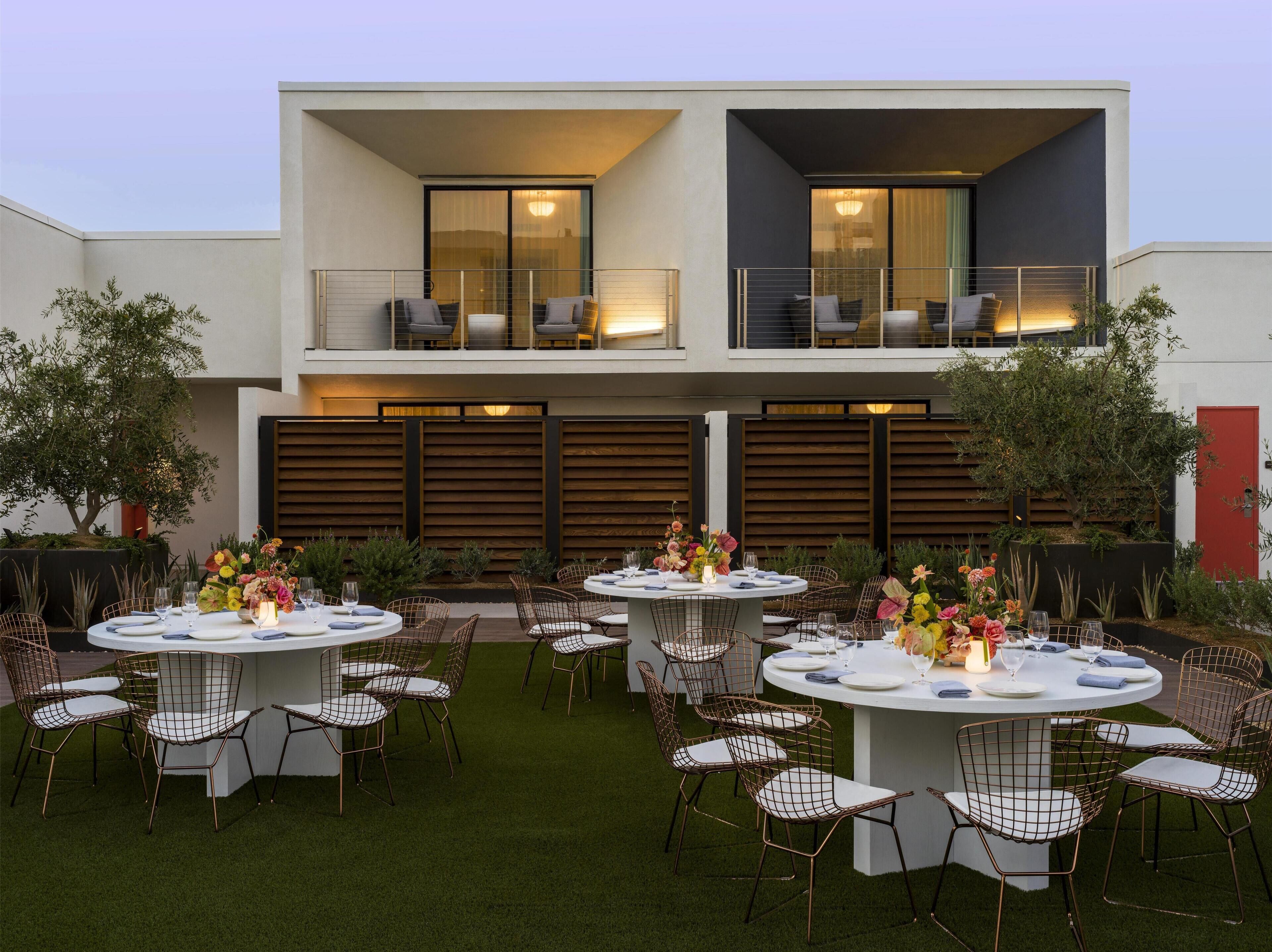 A modern outdoor dining area with round tables and wire mesh chairs, set against a building with balconies and a red door.