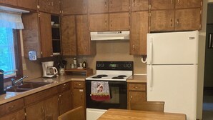 A compact kitchen with wooden cabinets, a stove, a refrigerator, and a dining table with chairs. - Charming 2-bedroom cottage in Fish Creek with AC, heat, WiFi (Fish Creek)