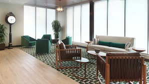 A modern lobby with a wooden floor, green and white patterned carpet, a large clock, green chairs, a beige sofa with a green pillow, and a large window with vertical blinds. - Fairfield By Marriott® Inn & Suites Grovetown (Grovetown)