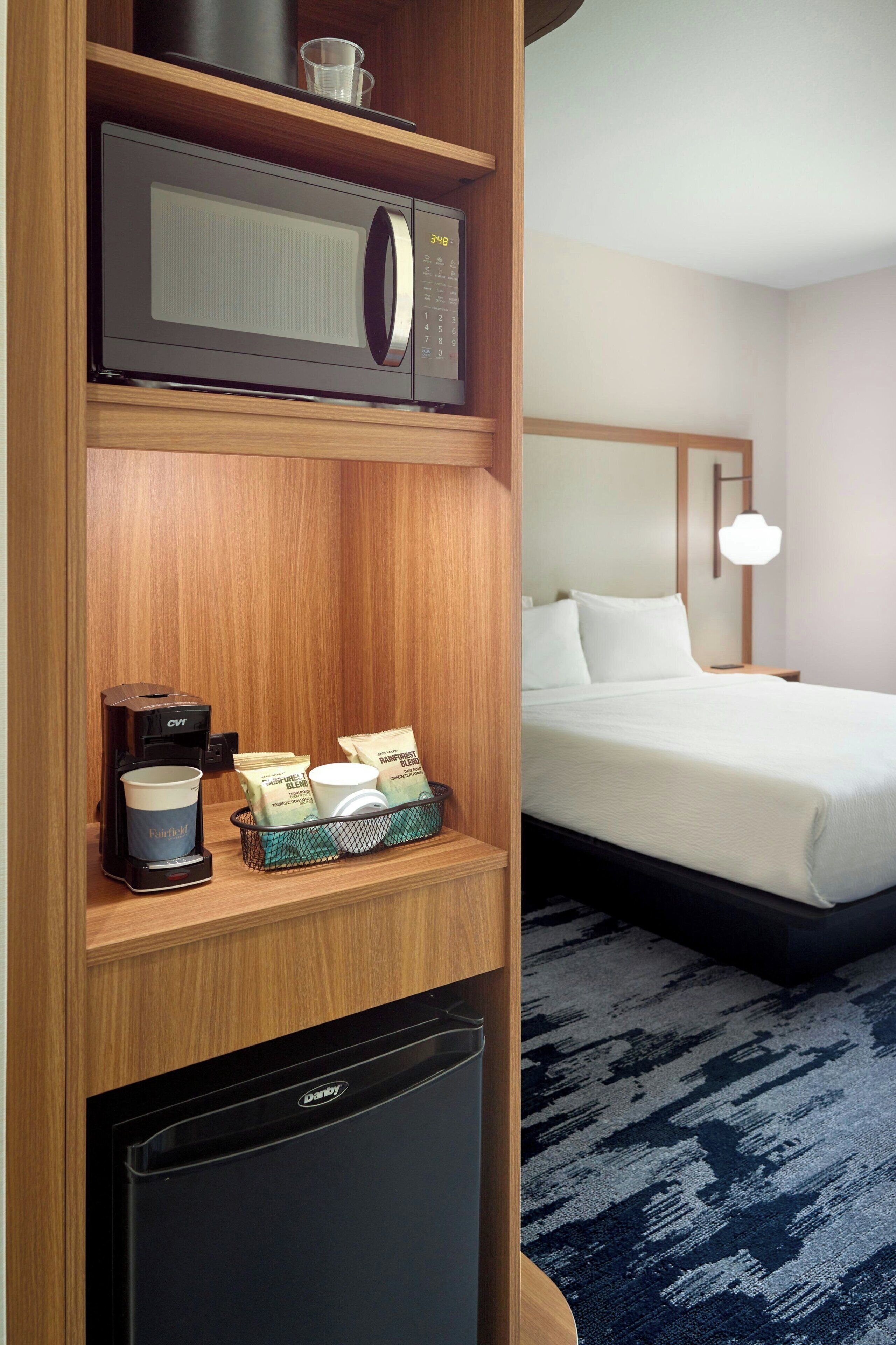 A hotel room with a microwave, coffee maker, and a mini fridge on the shelf, a bed with white linens, and a blue patterned carpet.