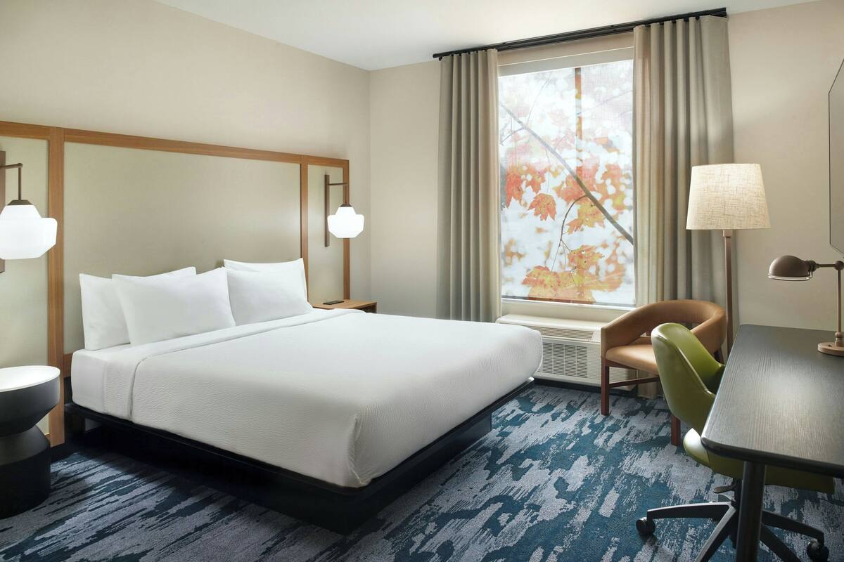 A hotel room with a large bed, a desk, a chair, a lamp, and a window with curtains.