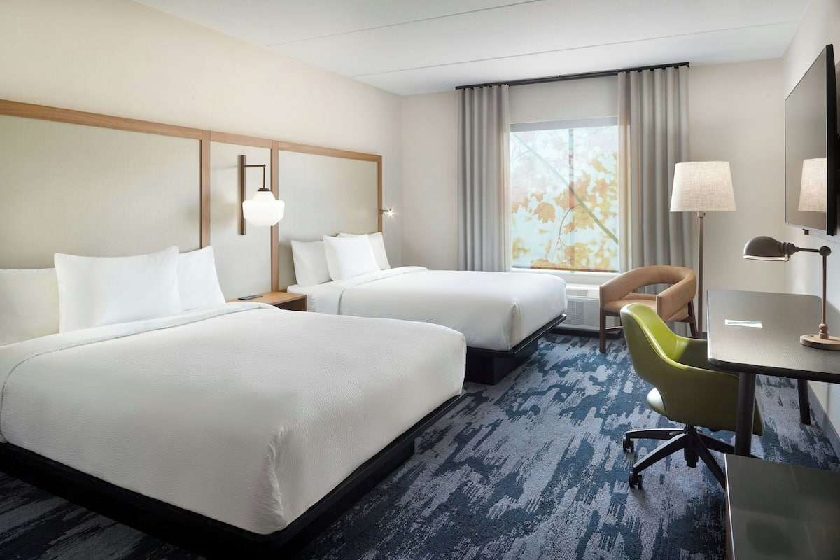 A hotel room with two beds, a desk, a chair, and a large window with curtains.