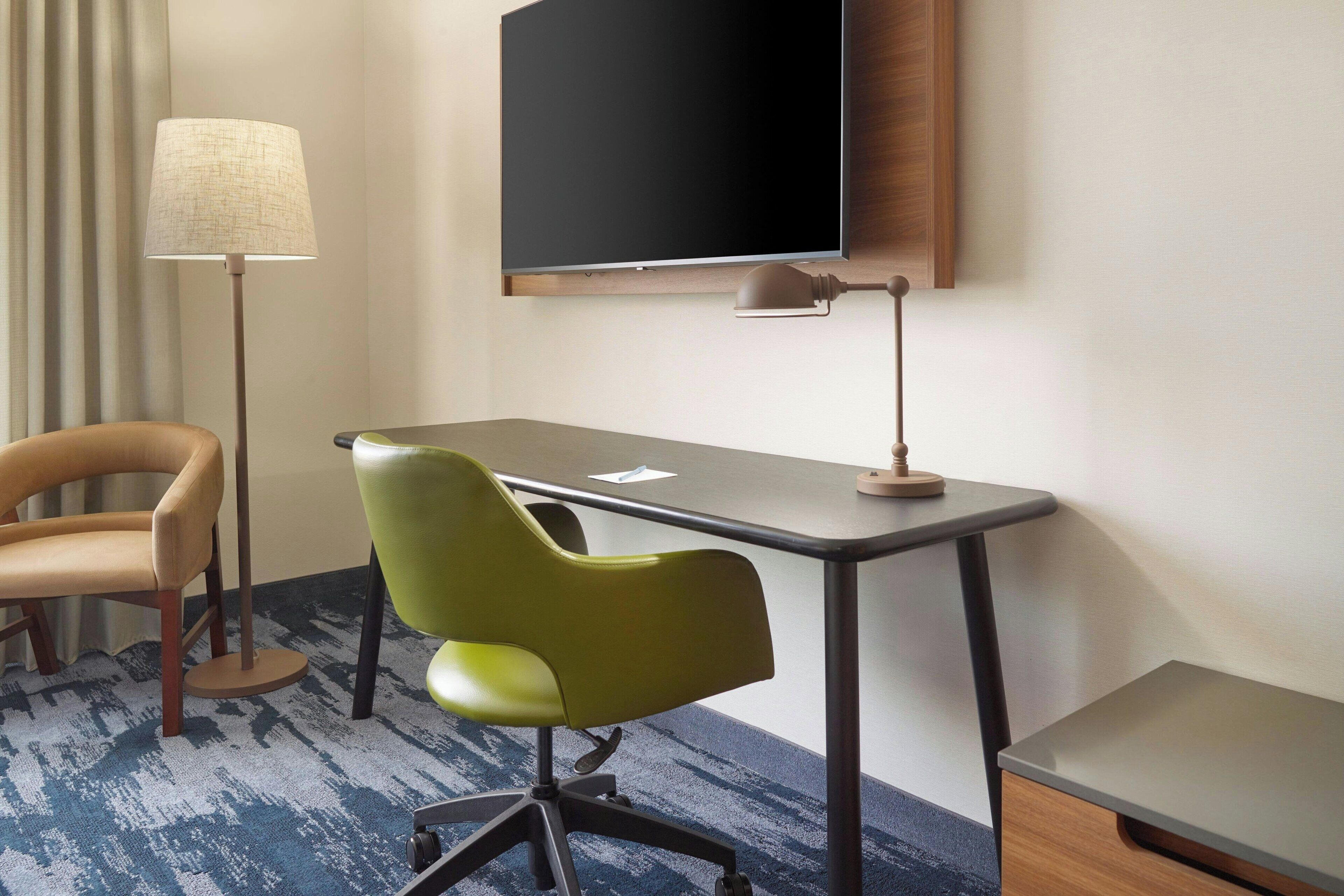 A hotel room with a desk, a green chair, a brown chair, a lamp, and a flat-screen TV mounted on the wall.