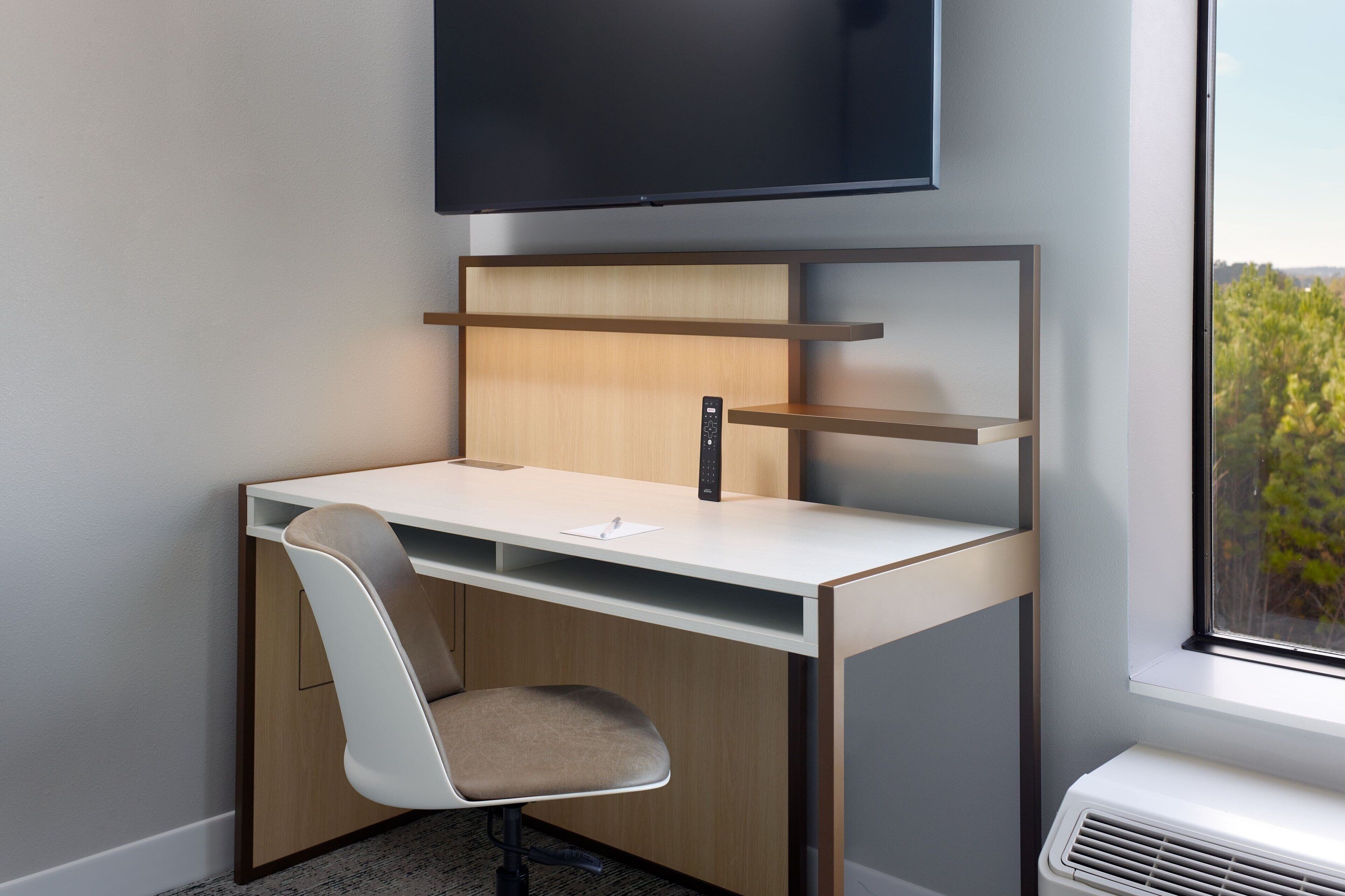 A modern office space with a desk, chair, mounted television, and a window with a view of greenery.
