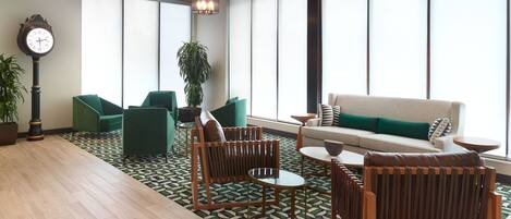 A modern lounge area with green chairs, a sofa, and a round table. A large clock is on the wall, and there are large windows providing natural light.