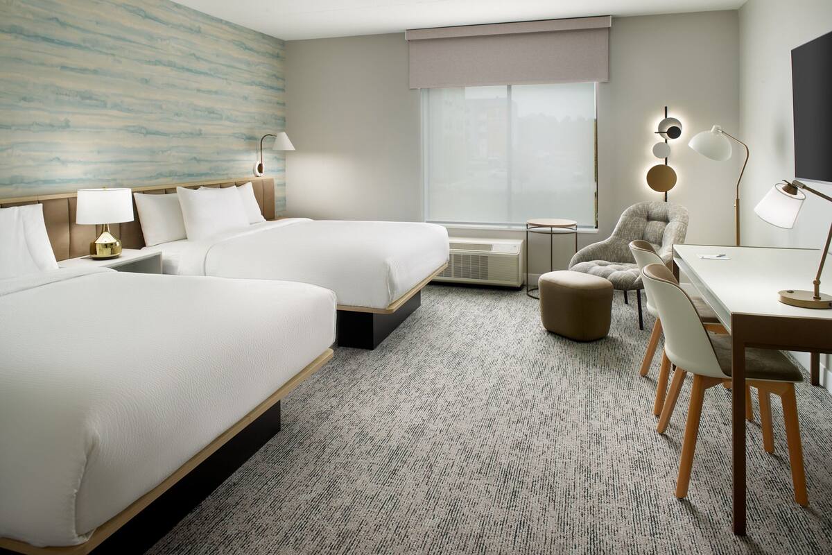 Une chambre d’hôtel avec deux lits, un bureau, une chaise, une lampe et un luminaire fixé au mur.