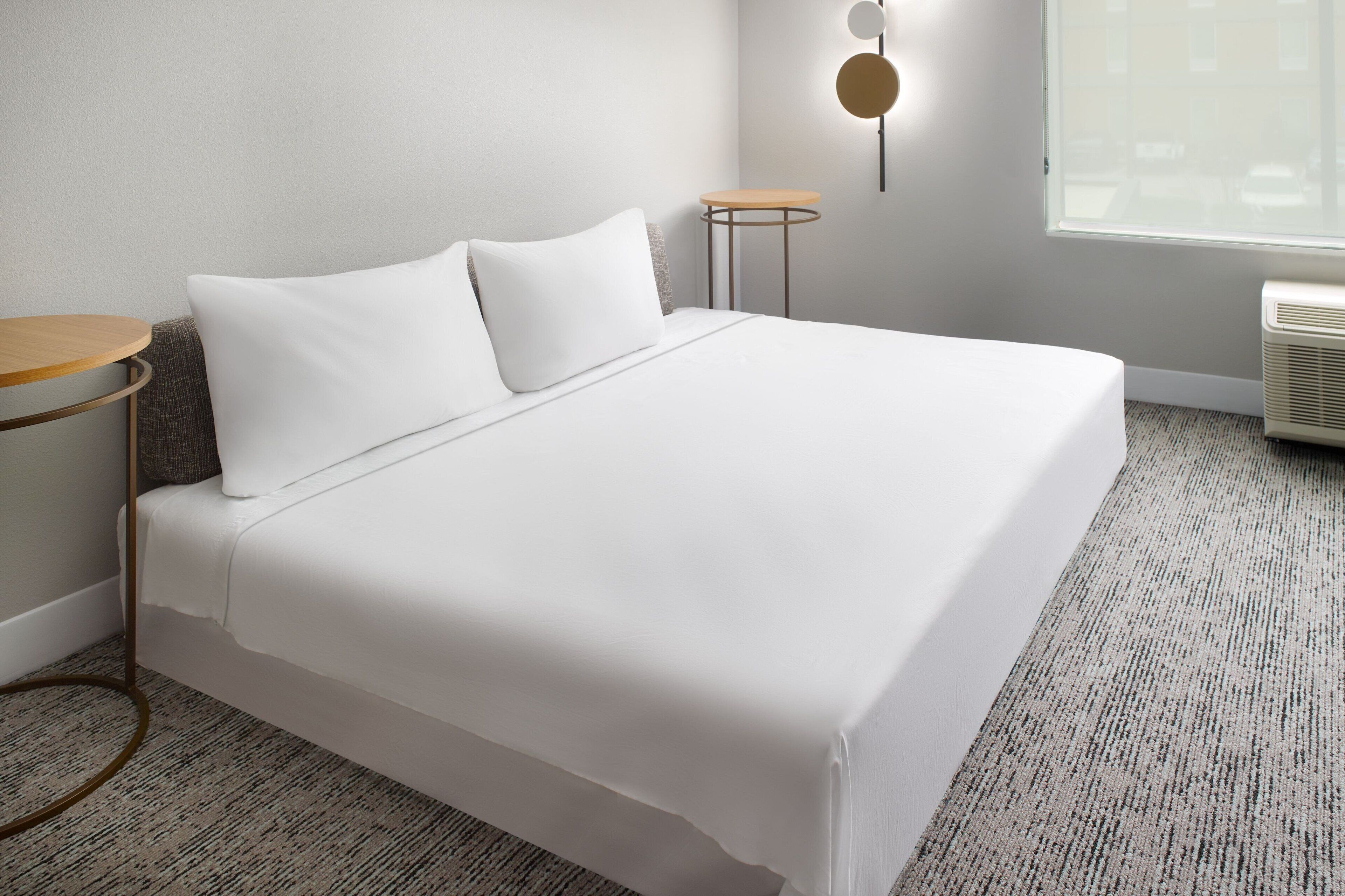 A neatly made bed with white linens, a small round table with a wooden top, and a wall-mounted light fixture.