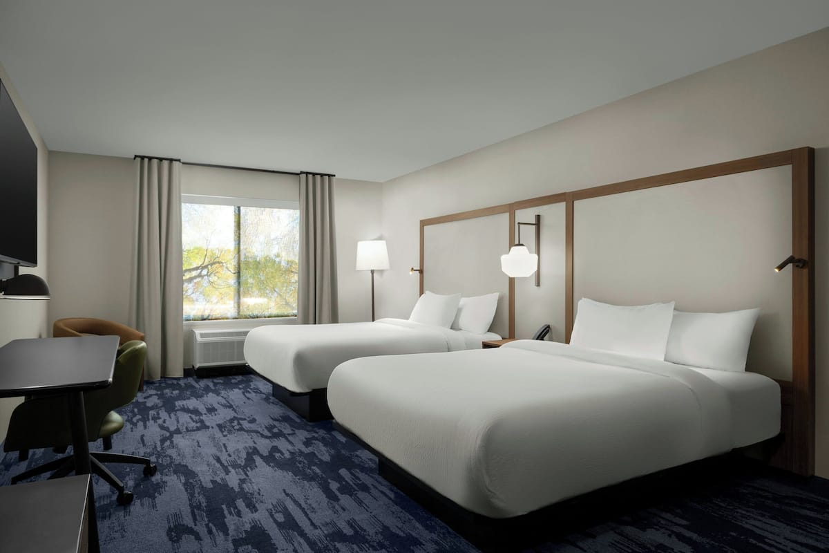 A hotel room with two beds, a desk, a chair, a TV, and a large window with curtains.