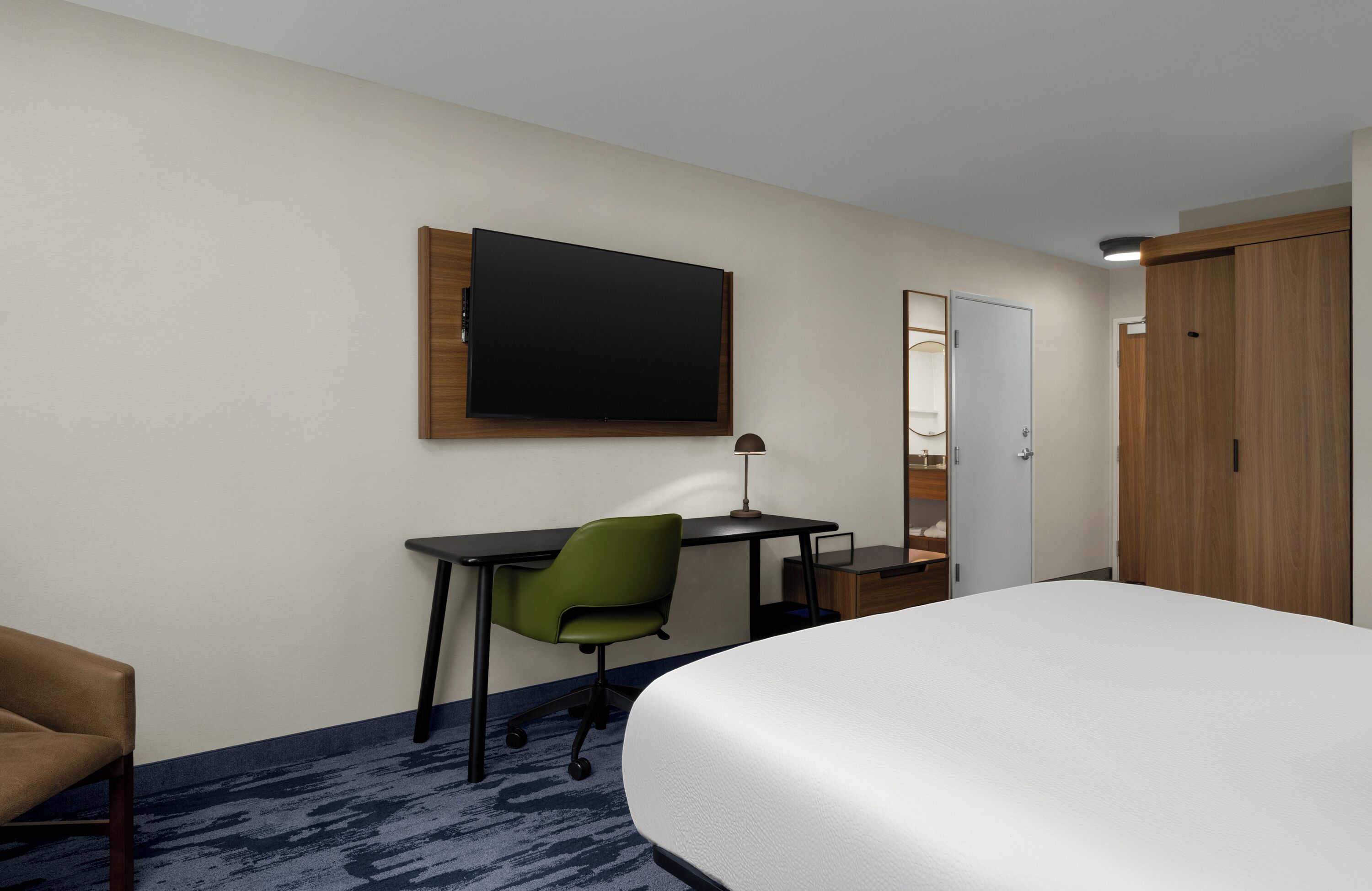 A hotel room with a bed, a desk with a chair, a flat-screen TV, a mirror, and a wardrobe.