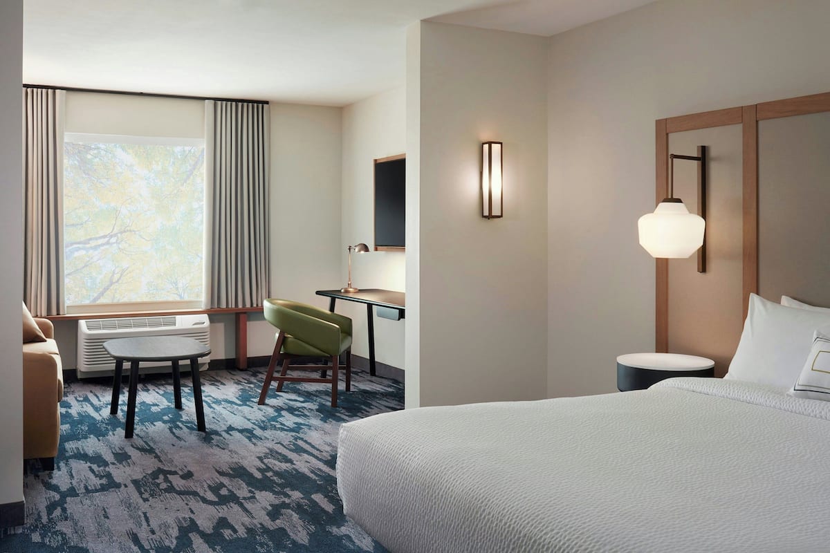 A hotel room with a bed, a desk, a chair, a small table, and a lamp.