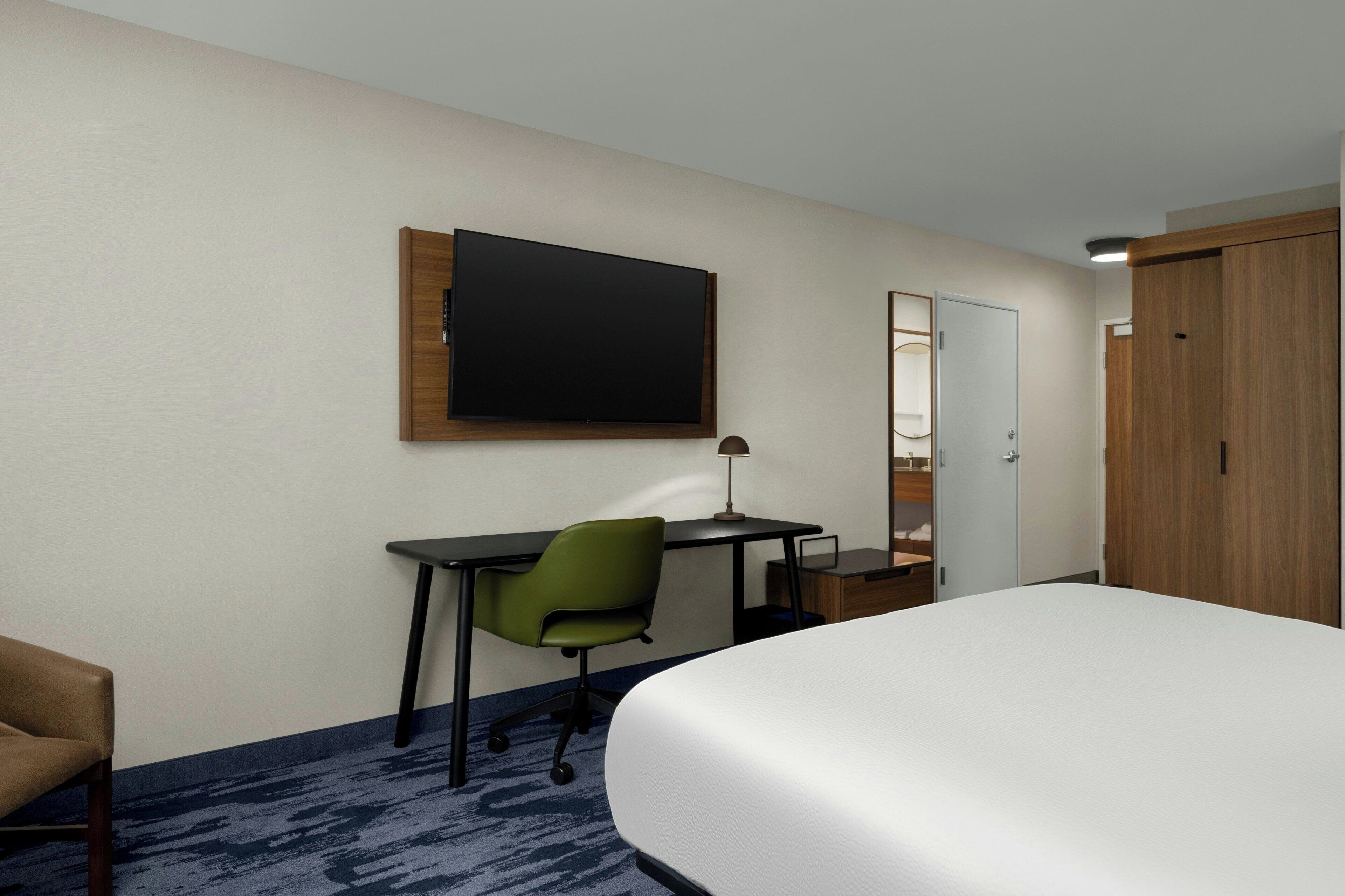 Quarto de hotel com cama, escrivaninha com cadeira, televisão de tela plana na parede e guarda-roupa.