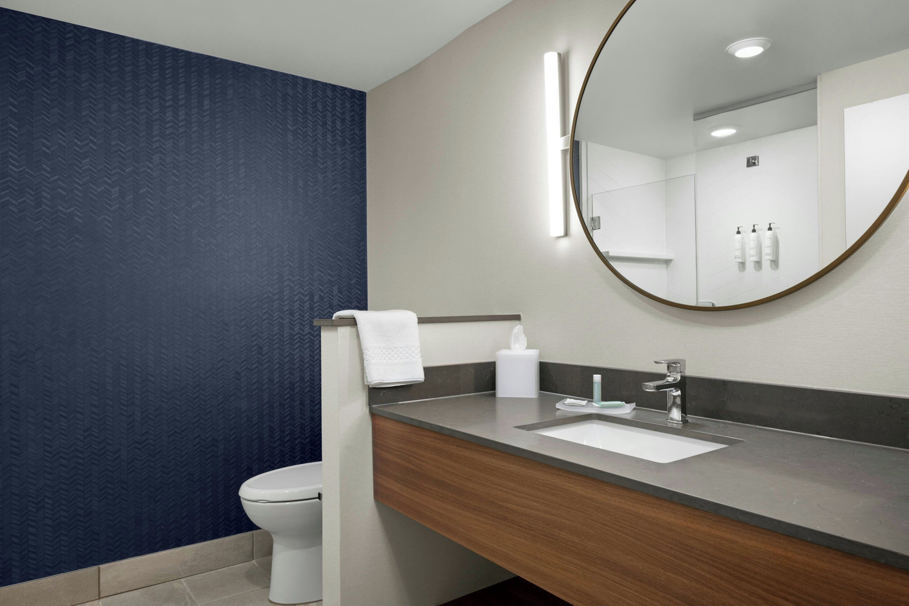 A modern bathroom with a white toilet, a wooden vanity, a large mirror, and a wall-mounted sink.
